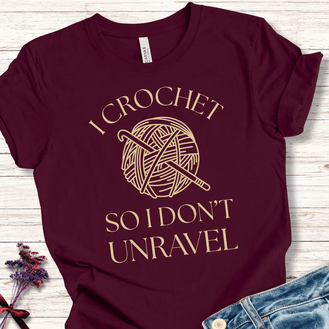 Maroon t-shirt with golden 'I Crochet So I Don't Unravel' text and yarn graphic, ideal for crochet lovers.