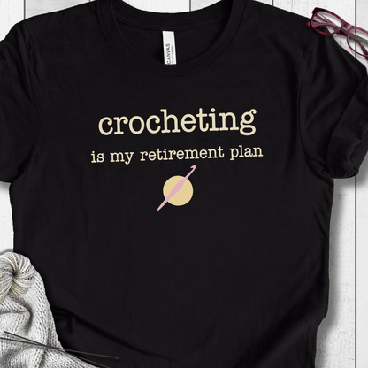 Black cotton t-shirt with 'crocheting is my retirement plan' slogan and crochet hook graphic.