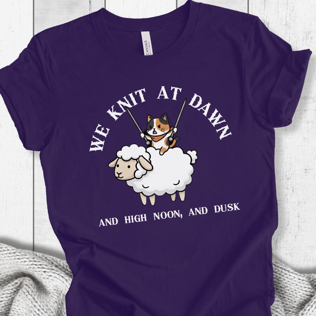 Purple t-shirt featuring a whimsical cartoon cat knitting on a sheep with the text 'We Knit at Dawn'.