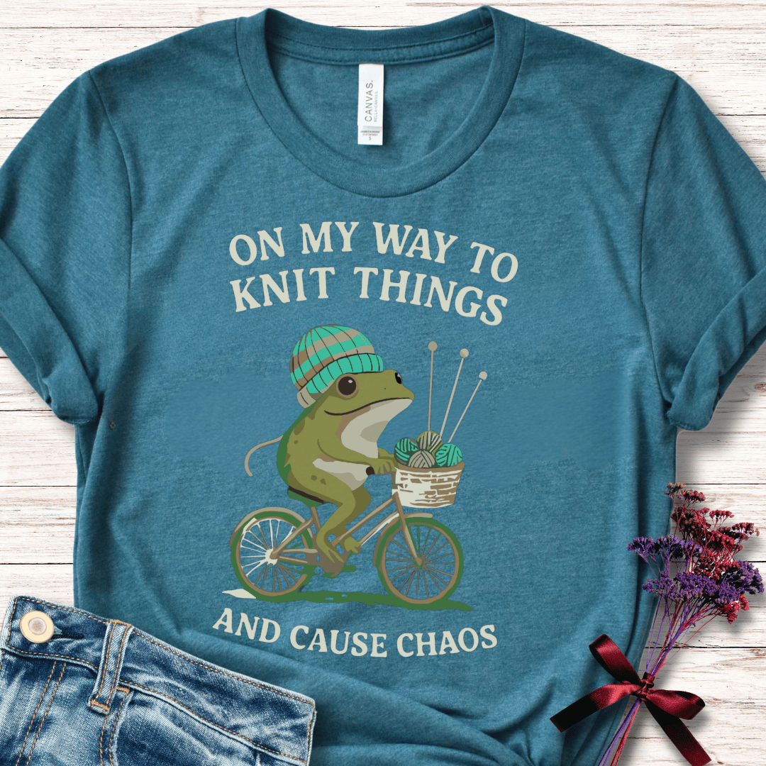 Teal t-shirt featuring a playful frog on a bicycle with knitting supplies, perfect for craft lovers.