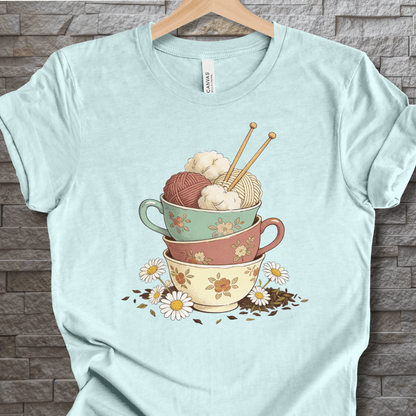 Light blue t-shirt featuring colorful yarn bowls and knitting needles design, perfect for craft lovers.