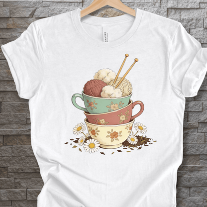 White t-shirt featuring colorful stacked cups with yarn and knitting needles, floral design.