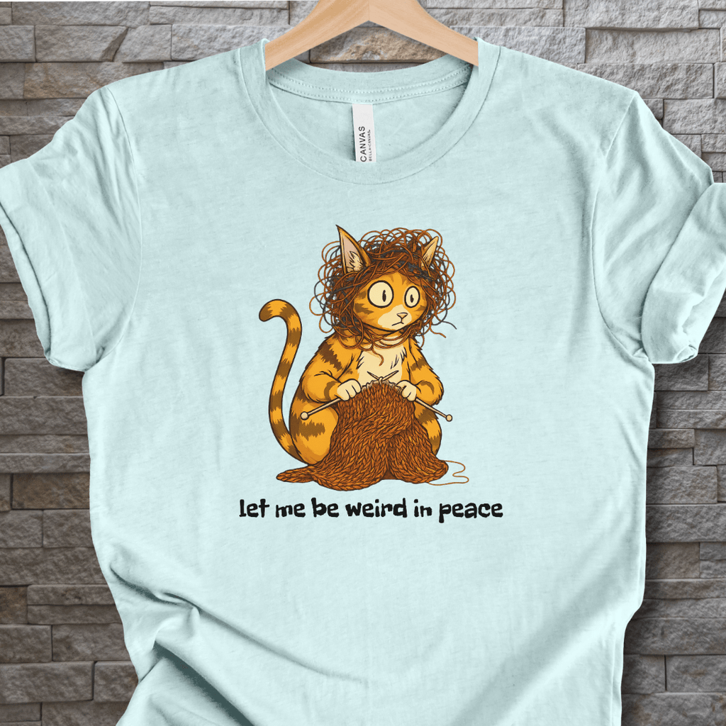 Funny cat graphic t-shirt in light blue, featuring a quirky cat knitting with the text 'let me be weird in peace'.