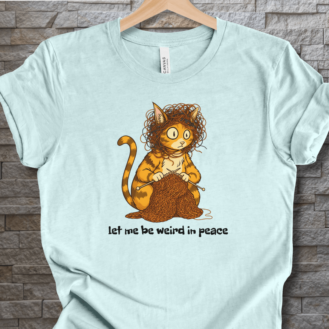 Funny cat graphic t-shirt in light blue, featuring a quirky cat knitting with the text 'let me be weird in peace'.