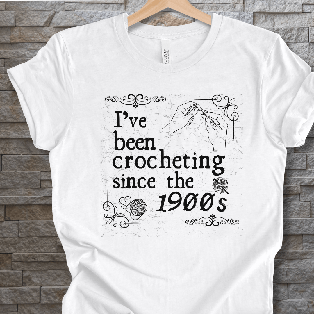 White t-shirt featuring crochet-themed graphic text 'I've been crocheting since the 1900s'.